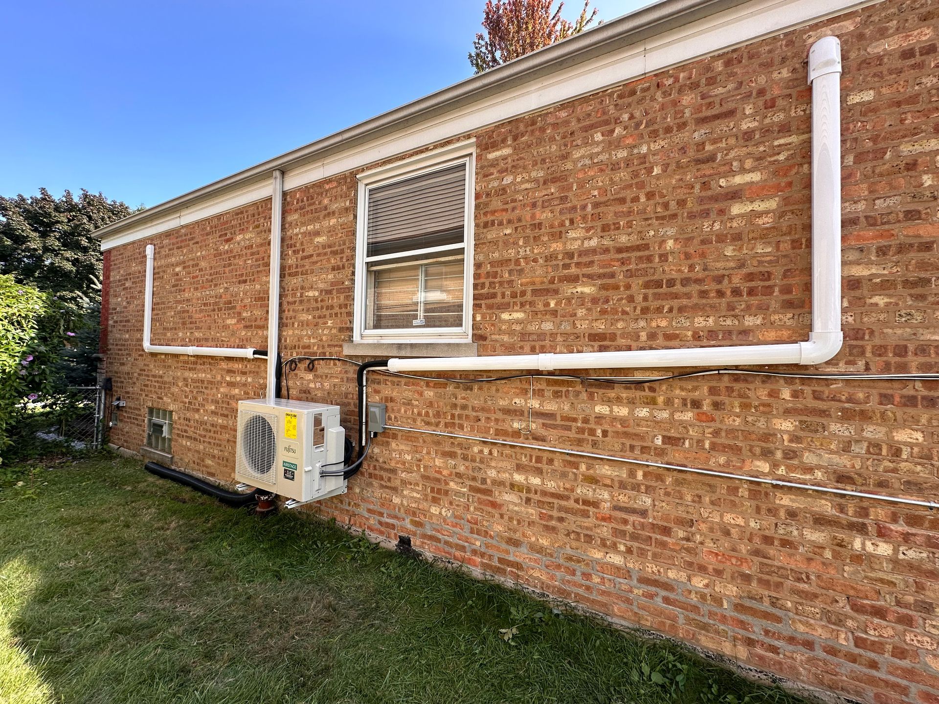 Brick house side wall with white pipes, window, and exterior air-conditioning unit on the lawn