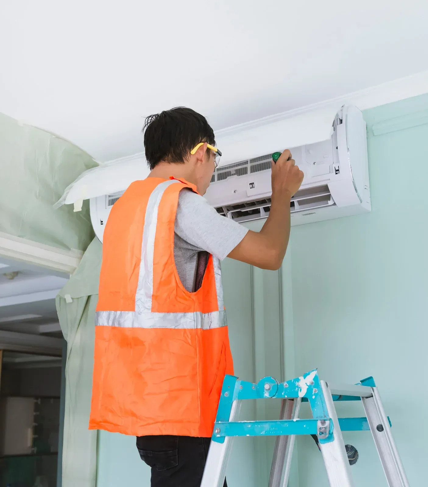 Technician in orange vest repairing a wall-mounted air conditioner on a stepladder indoors