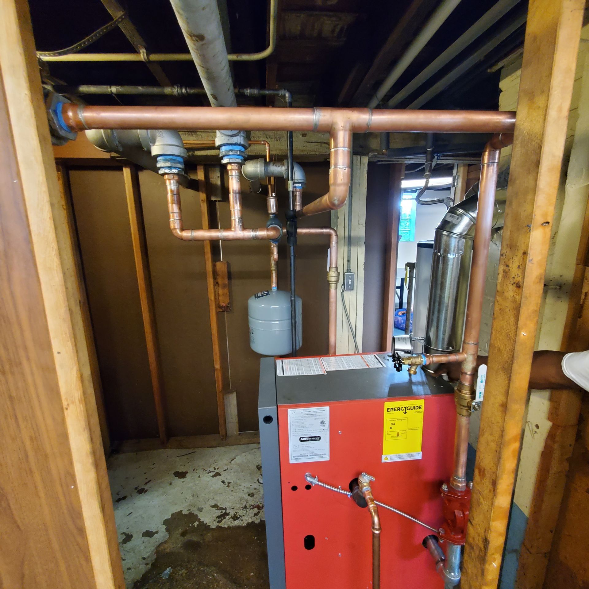 Red boiler room with copper pipes, gray tank, and a red furnace unit in a wooden-framed basement.