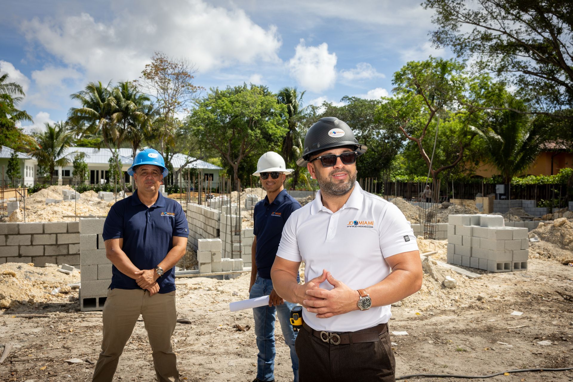 Three construction workers standing on a construction site with cement blocks. Two wear hard hats. Sunny outdoor scene.