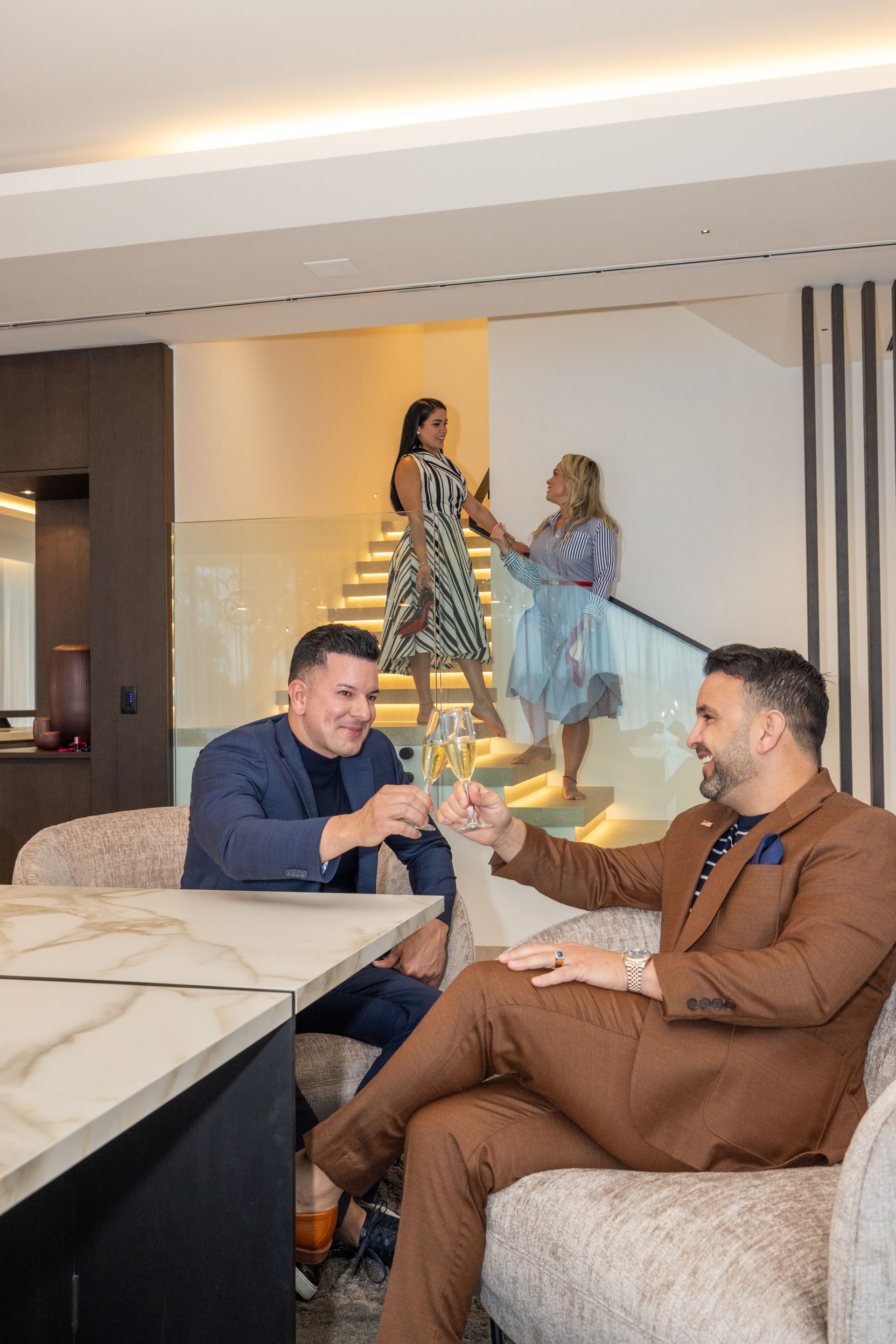 Two men toasting with champagne in a modern home. Two women are on the stairs behind them.