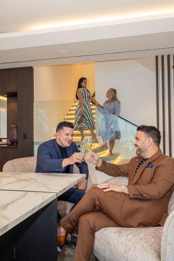 Two men toasting with champagne in a modern home. Two women are on the stairs behind them.