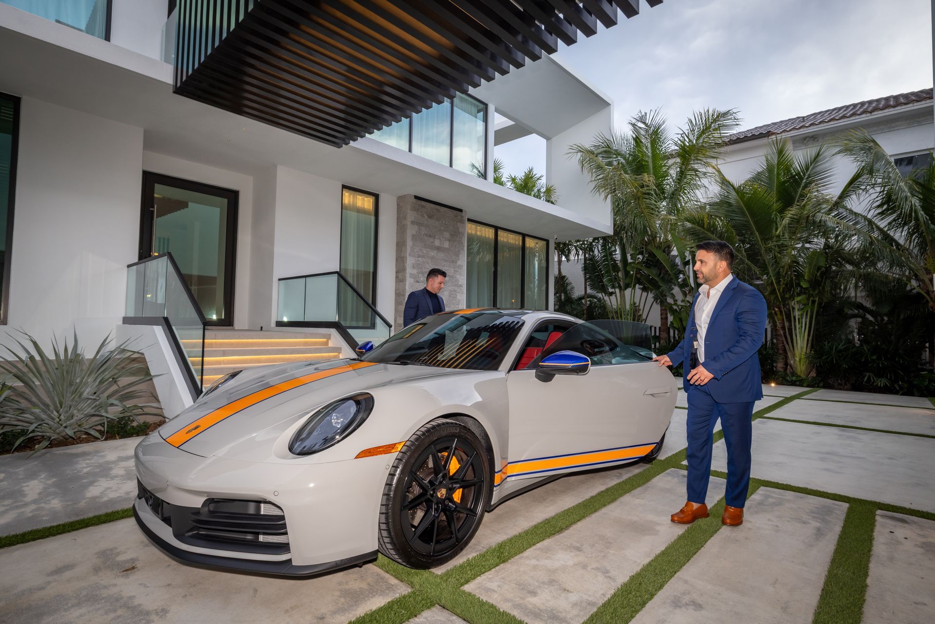 Man in suit next to a light gray Porsche with orange stripes parked in front of a modern white house.