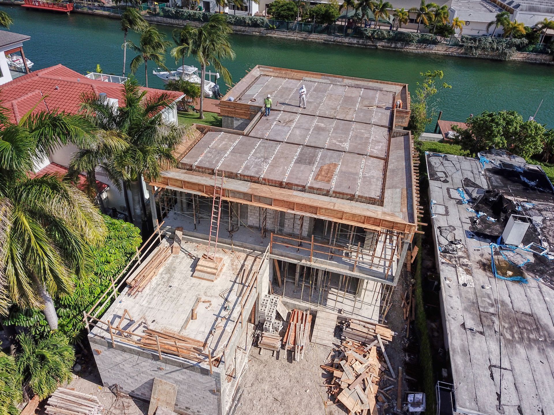 Construction site with two-story building frame near a canal. Workers on top level, surrounded by wood supports and materials.