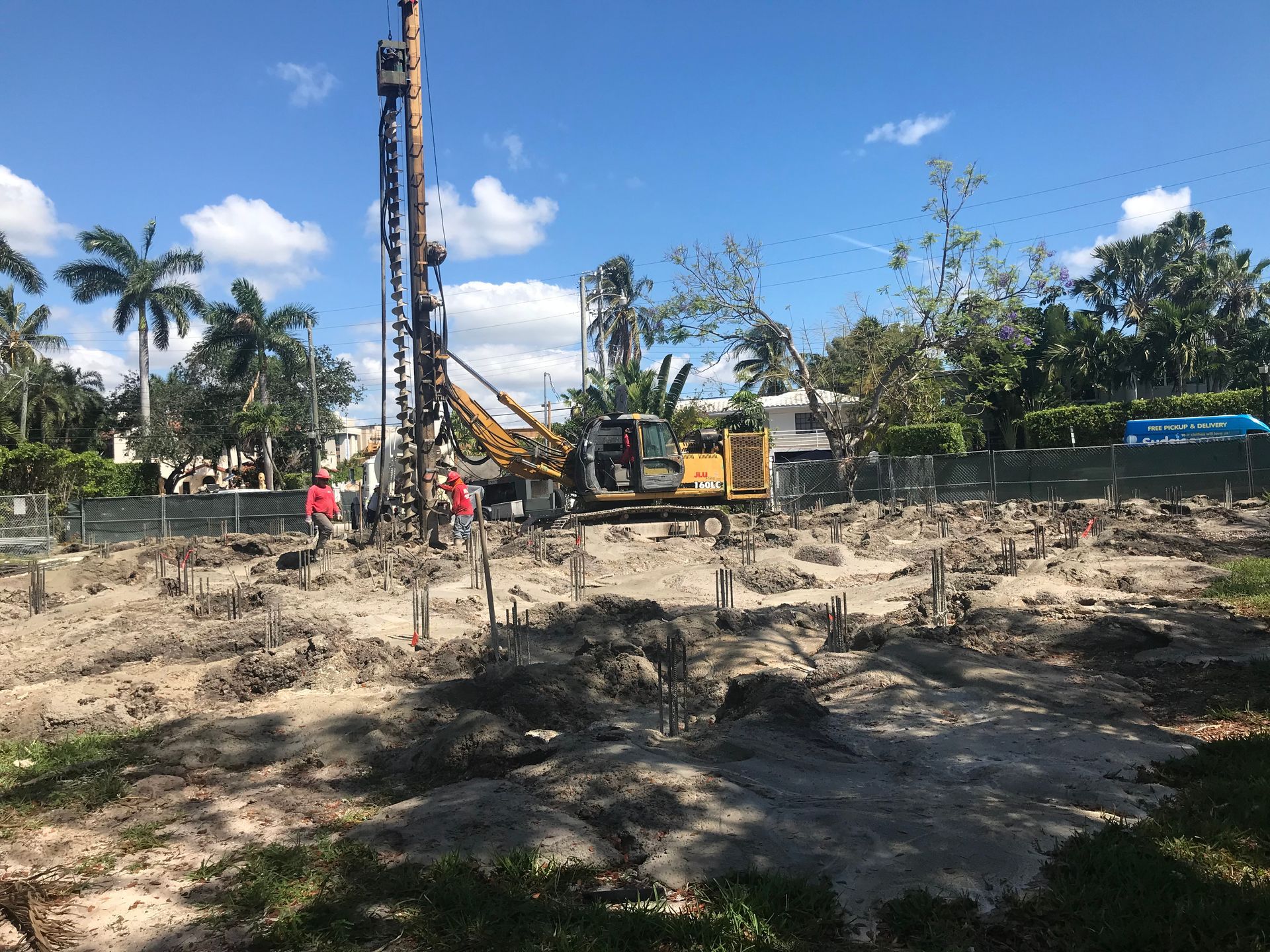 Construction site with pile driving machinery on dirt lot, palm trees in background.