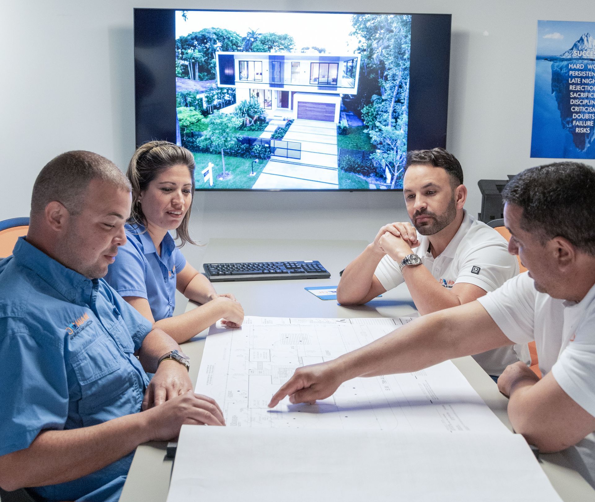 Four people reviewing blueprints around a table, with a house rendering on a screen in the background.