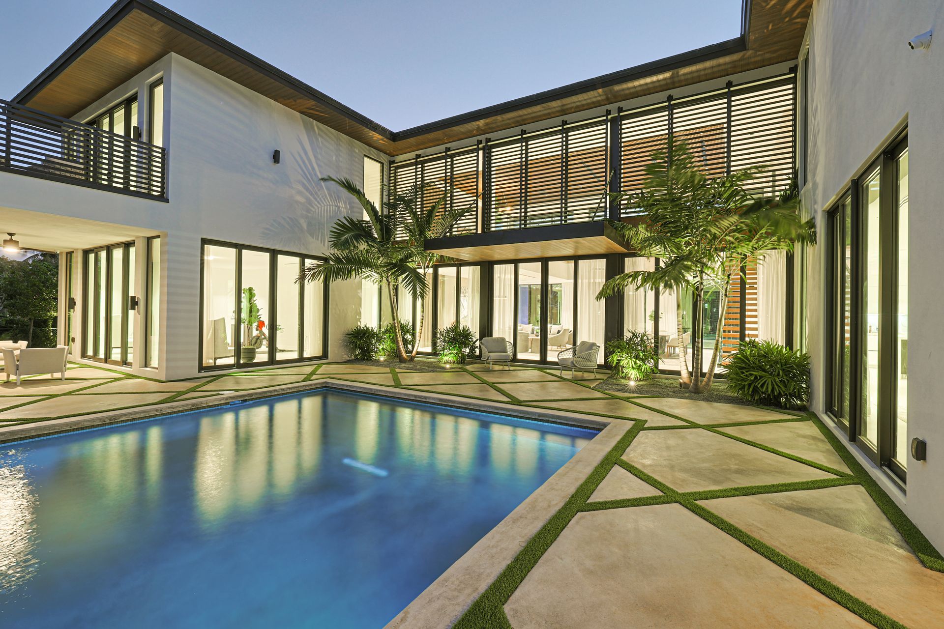 Pool in courtyard of a modern white house with large windows, illuminated at dusk.