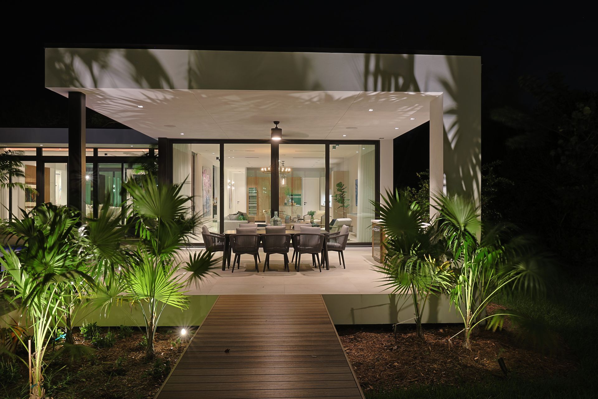 Lit patio with dining table at night, pathway leading up, plants in front, modern house in background.