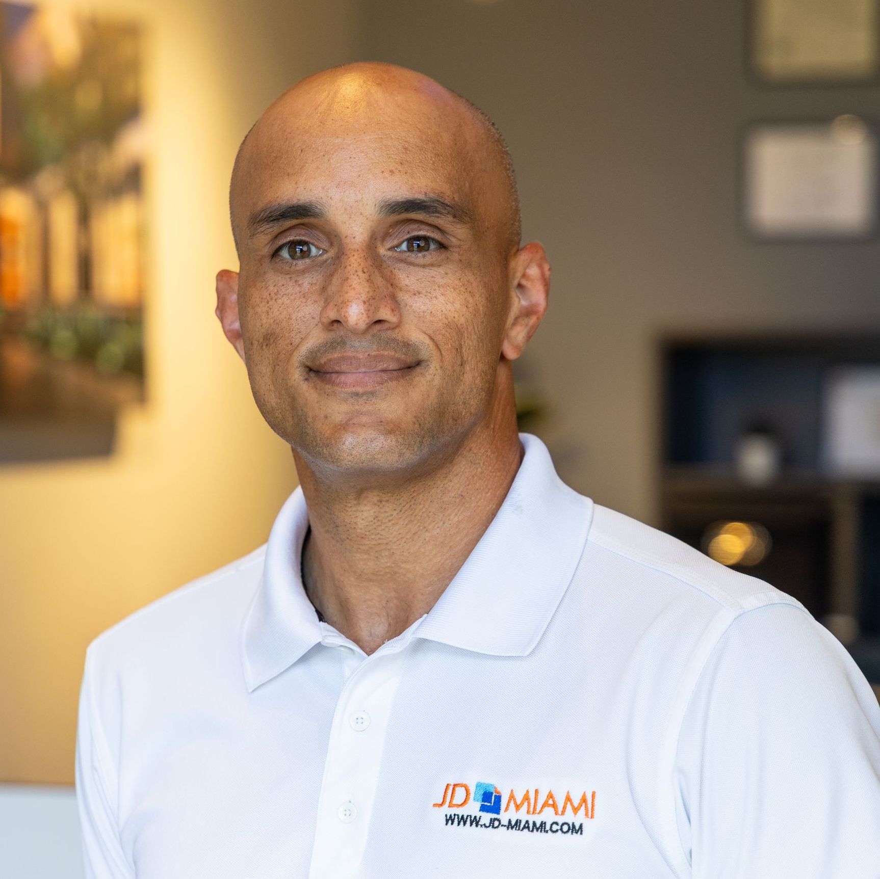 Man with shaved head wearing a white polo shirt smiles in an office setting.