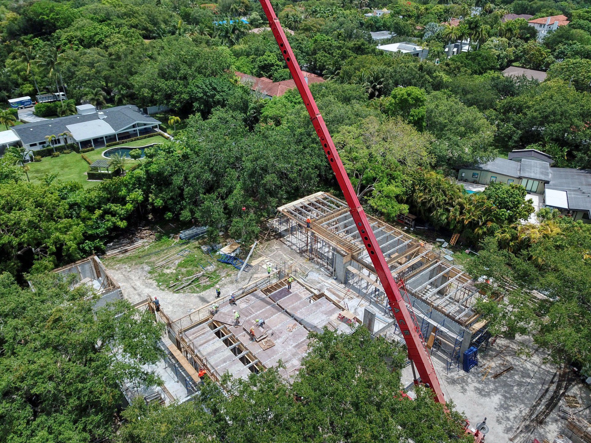 Construction site with red crane lifting materials, surrounded by trees and houses.