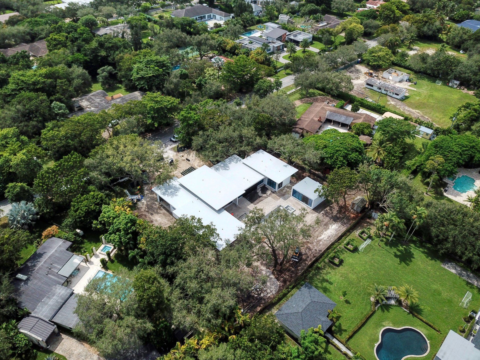 Aerial view of a home with a light-colored roof surrounded by trees and residential properties.