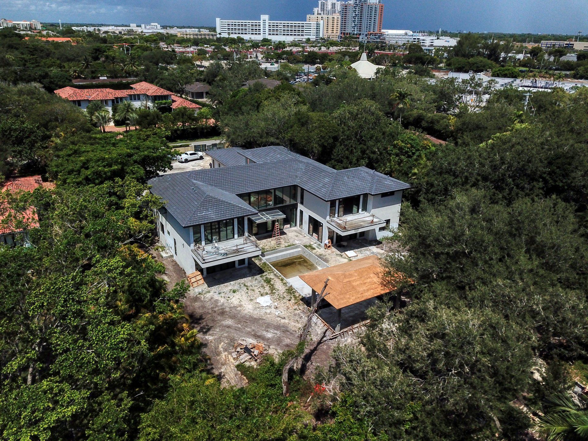 Aerial view of a gray modern house under construction, surrounded by lush green trees, with a building in the background.