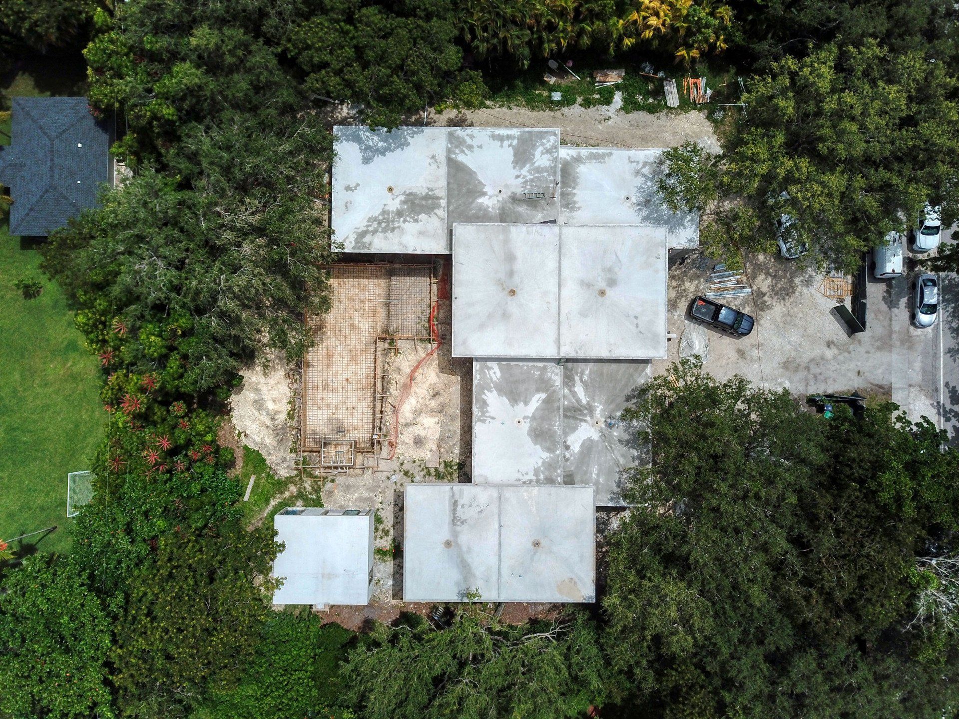 Aerial view of a building with visible roof sections surrounded by trees; a nearby gravel parking lot and green lawn are present.