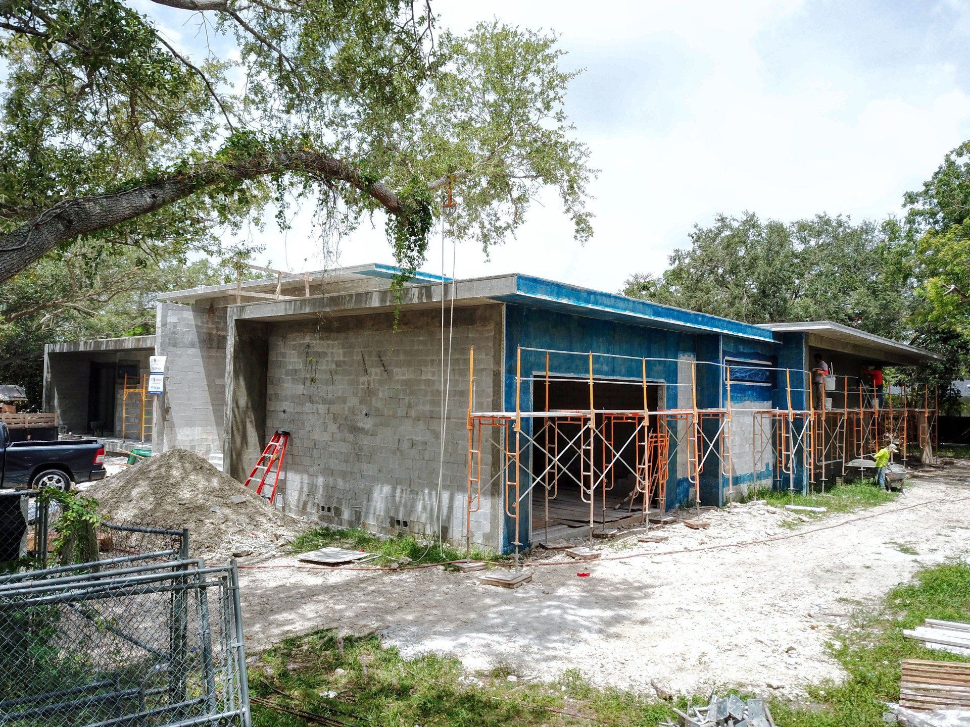 Building under construction; exterior view of a low-rise structure, scaffolding, dirt lot, blue siding, trees.