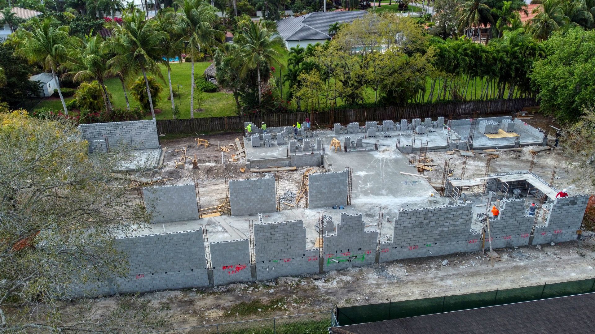 Construction site with concrete walls and workers, surrounded by trees and residential buildings.