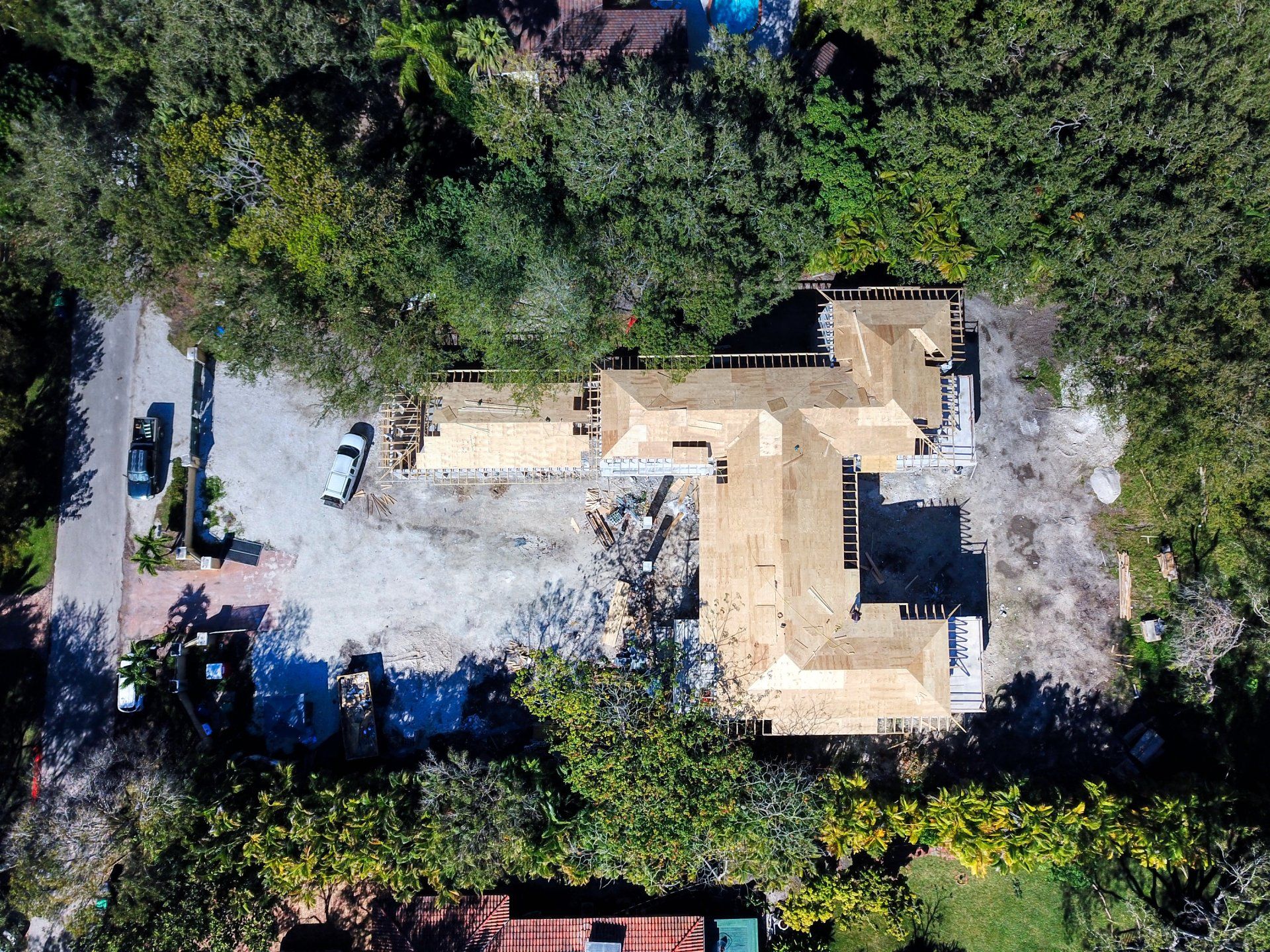 Aerial view of a home under construction with a light-colored roof, surrounded by trees and a gravel driveway.