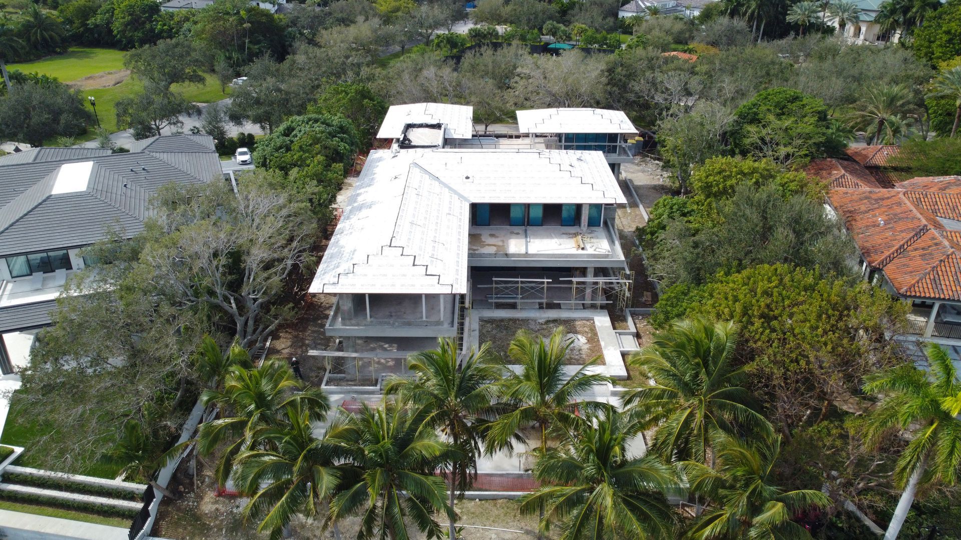 Aerial view of a modern white house with a flat roof, surrounded by green trees and palm trees.