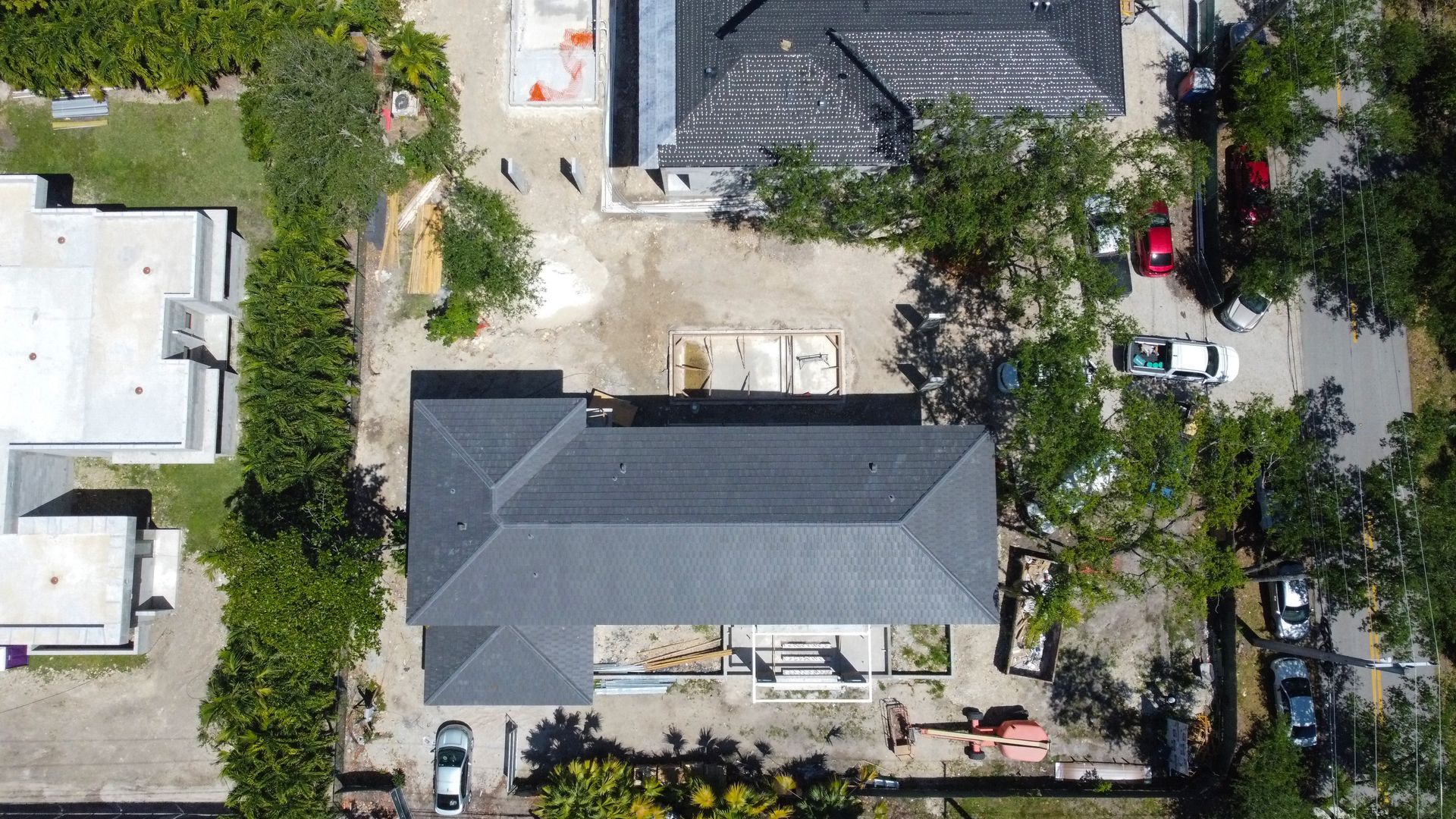 Overhead view of a building with a dark gray roof, surrounded by trees and construction materials on a sandy lot.