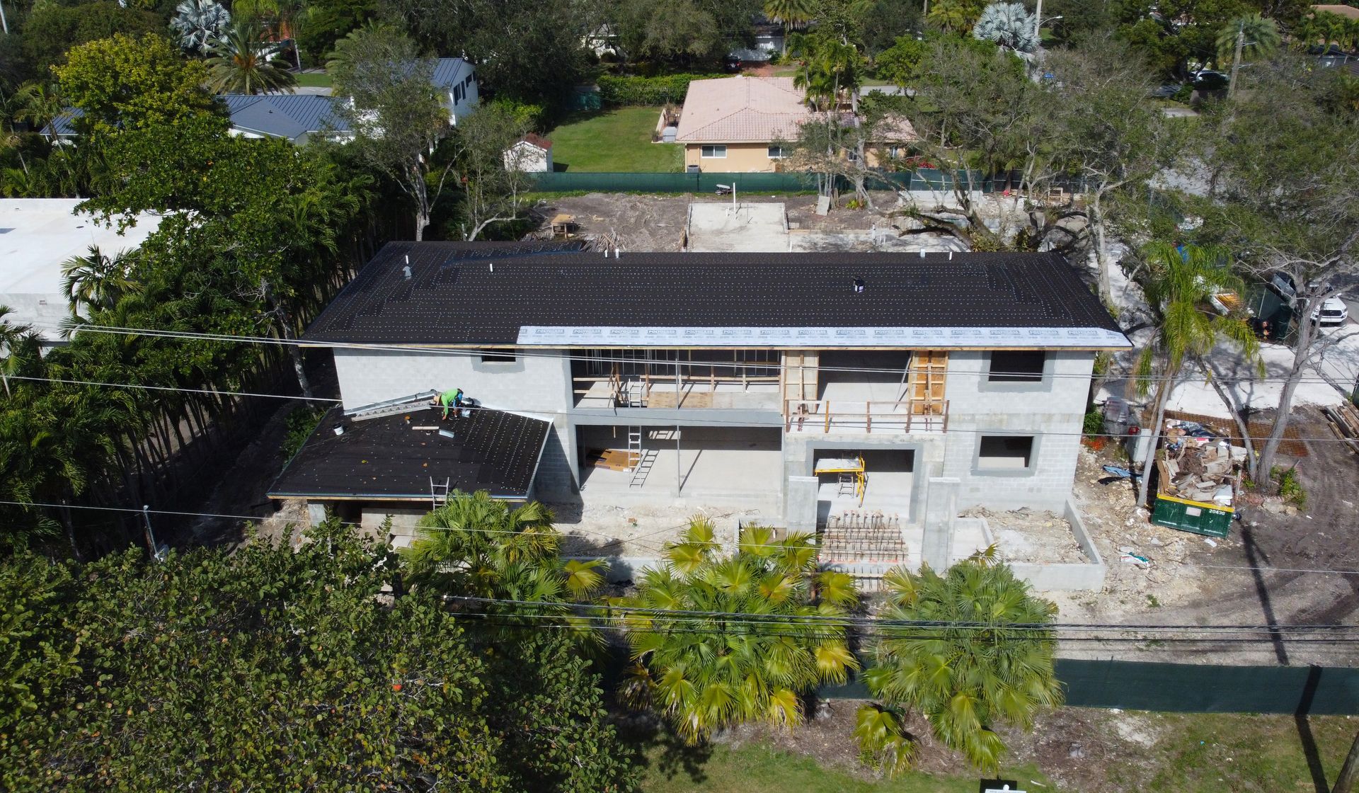 Aerial view of a two-story house under construction, with a partially completed roof and exposed framework, surrounded by trees.