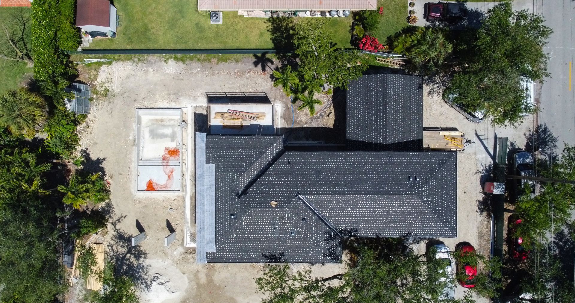 Aerial view of a building with a damaged black roof, construction site beside it, and surrounding trees.