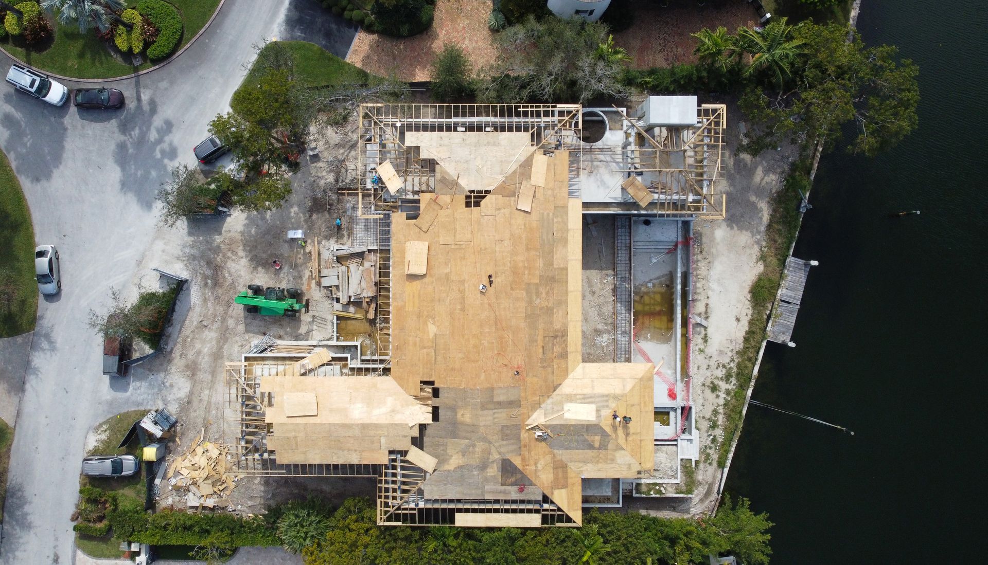 Aerial view of house under construction; wood framing and roofing, water and street visible.