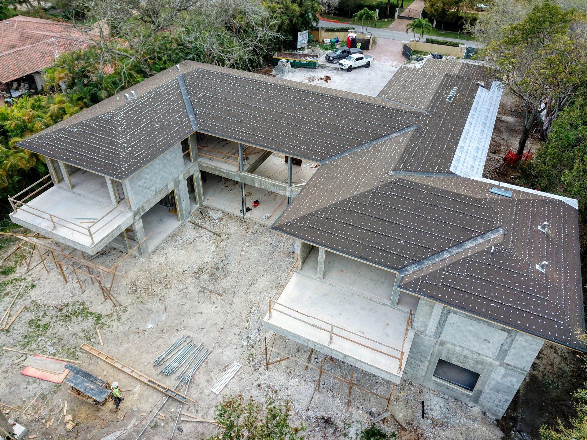 Aerial view of a house under construction with a gray roof and concrete structure; surrounded by trees and a dirt lot.
