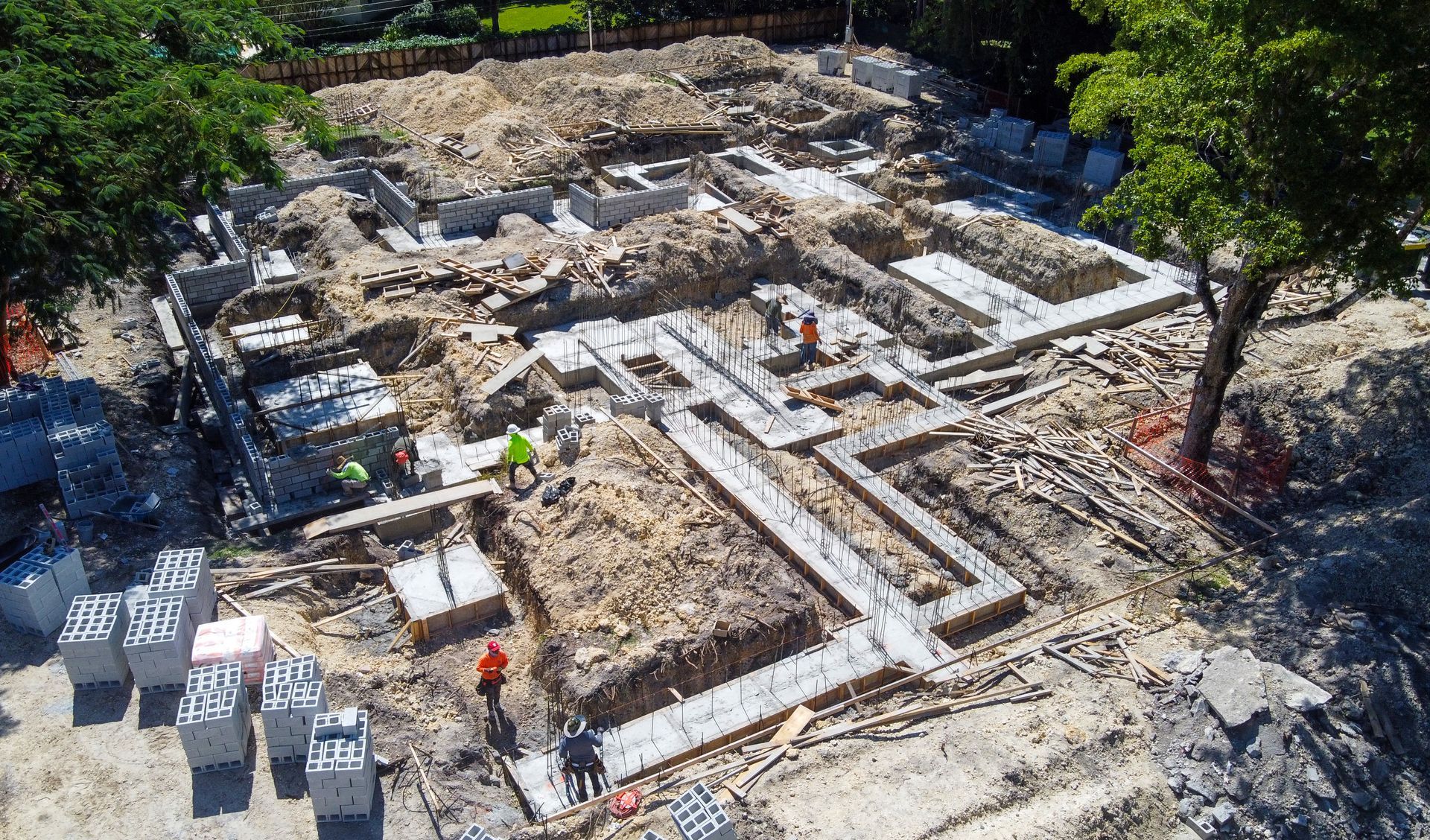 Construction site, foundation, concrete blocks, workers. Outdoor setting with dirt and trees.
