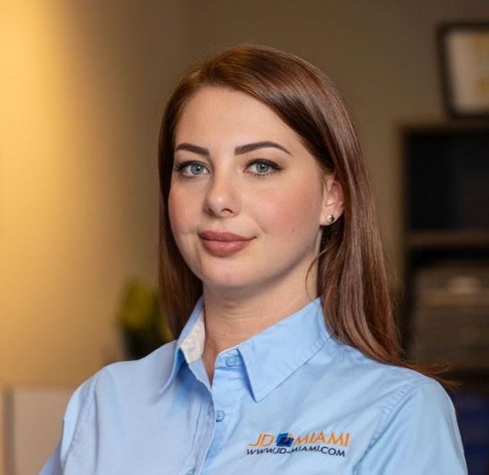 Woman with reddish-brown hair, wearing a blue shirt with a logo, smiling, indoors.