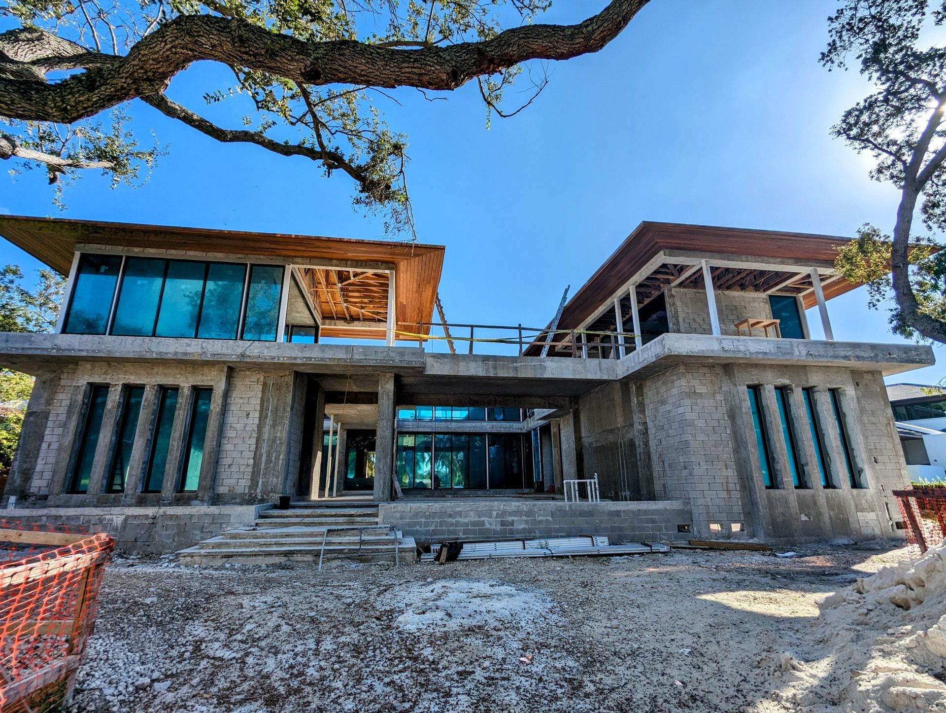 Two-story modern house under construction with large windows and a partially completed roof, set against a bright blue sky.