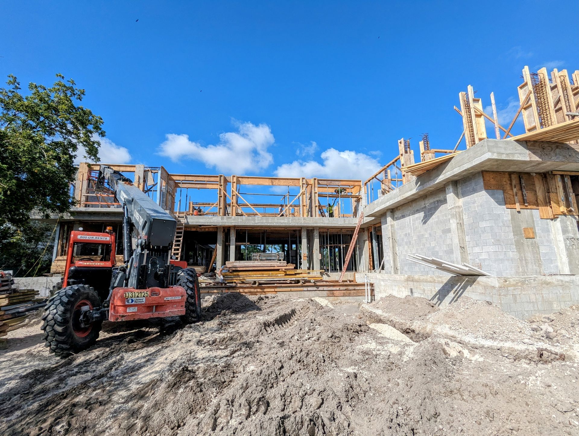 Construction site: framed house with concrete walls, heavy machinery in foreground, bright blue sky.
