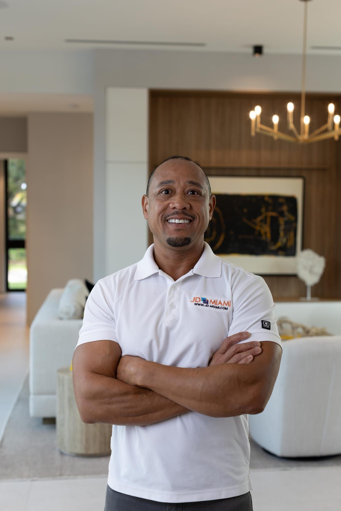 Man in white polo with arms crossed, standing in a modern living room.