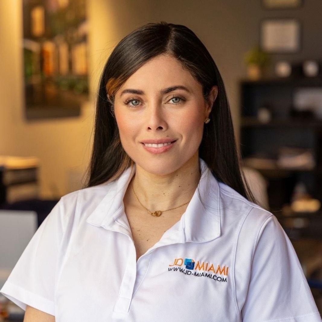 Woman with long dark hair, wearing a white shirt with an office background, smiling at the camera.