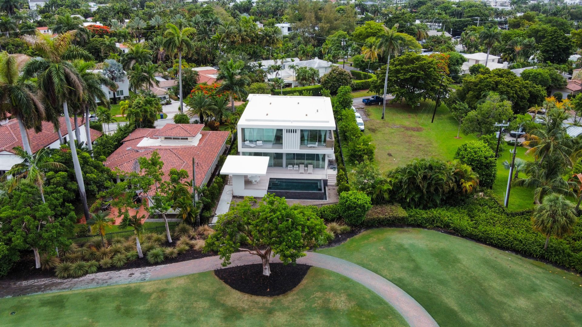Modern white house with a pool, next to a house with a red-tiled roof, surrounded by green trees and grass.