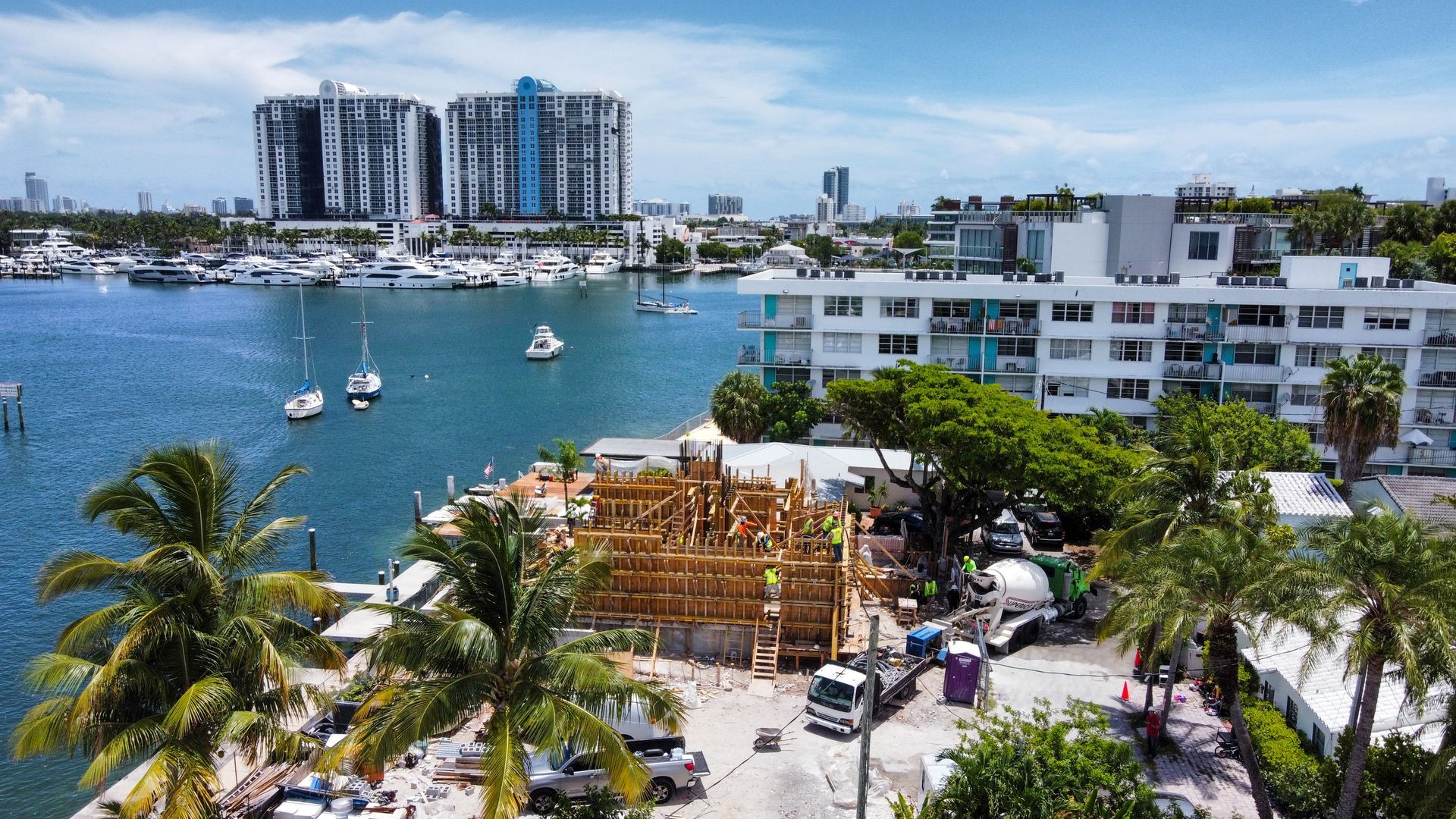 Construction site near water with buildings and boats; blue sky.