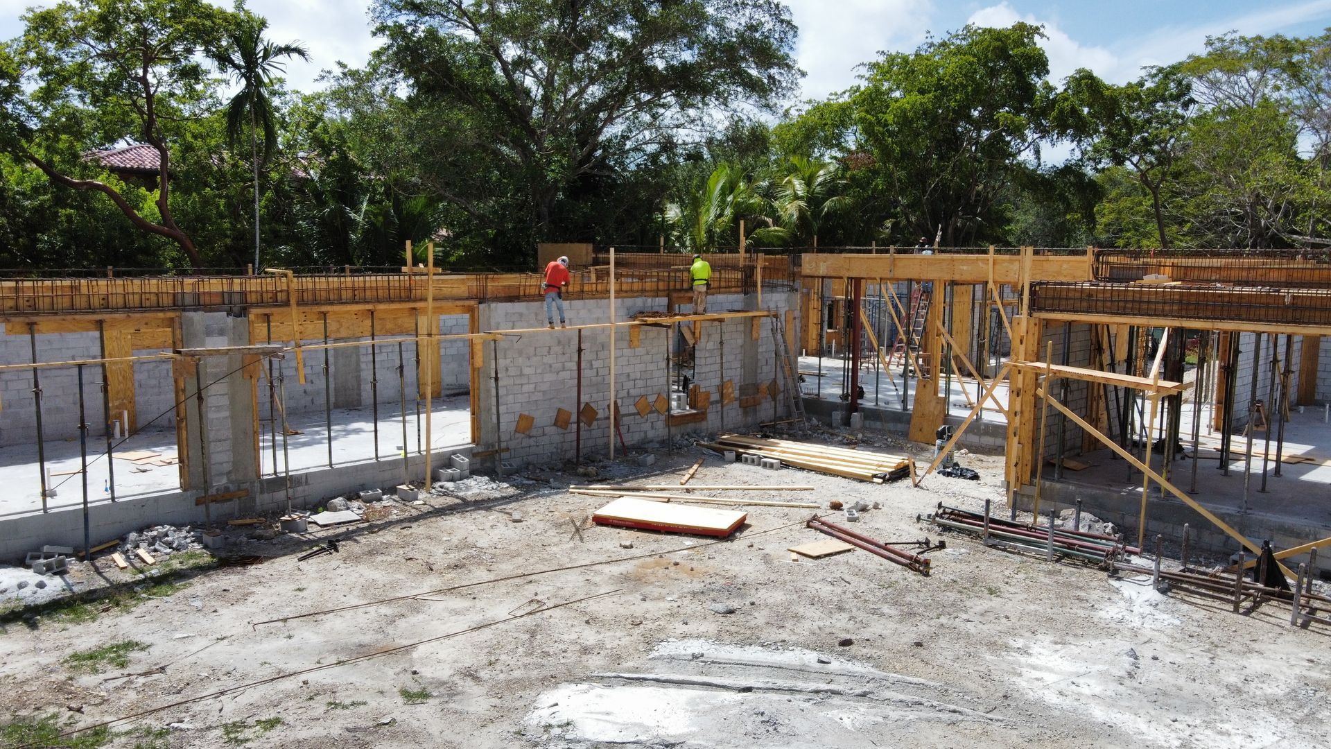 Construction site: Foundation walls and wooden framing, two workers in safety vests, surrounded by trees.