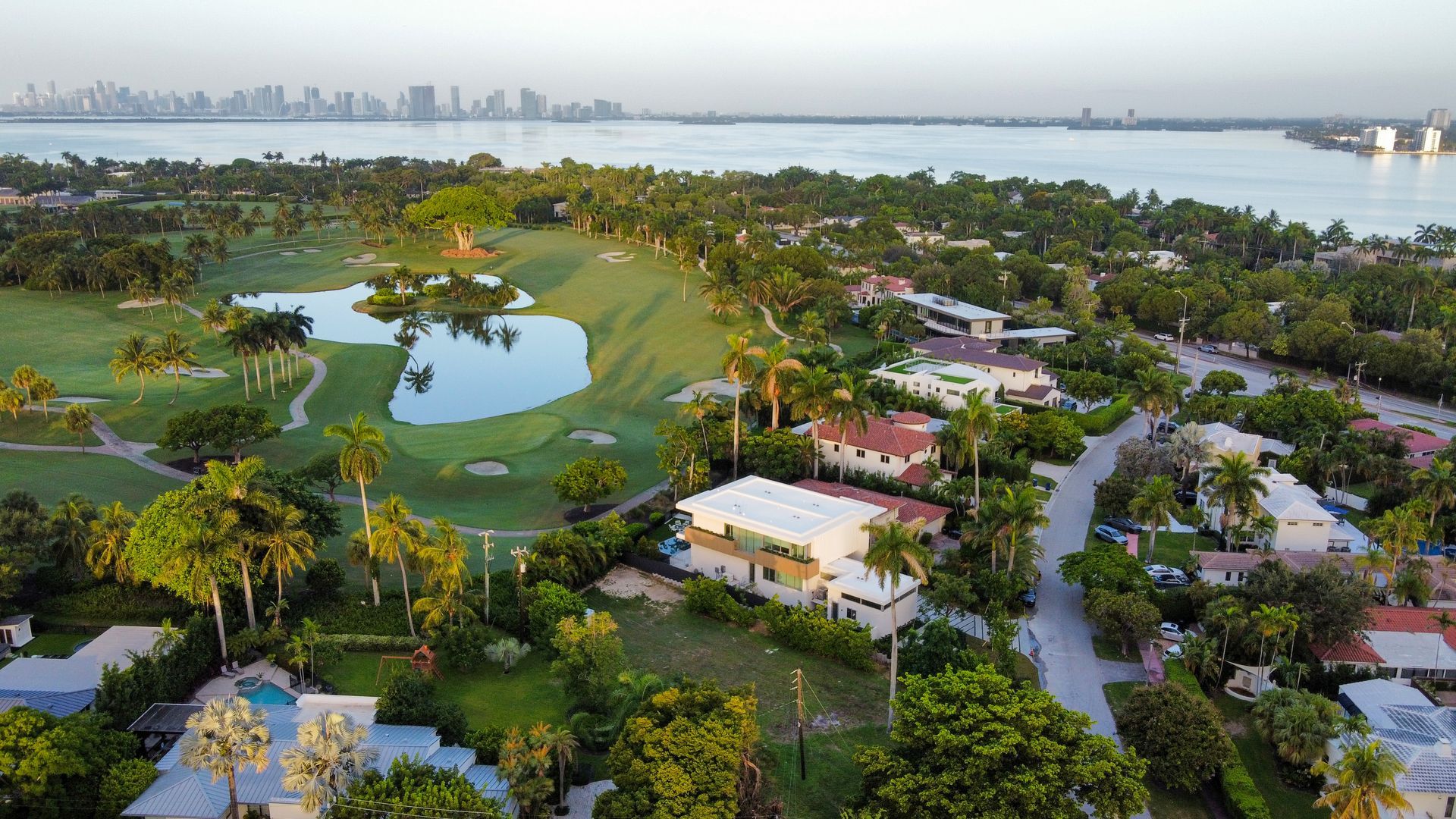 Aerial view: golf course with lake and homes, city skyline in background. Green trees, blue water, sunny.