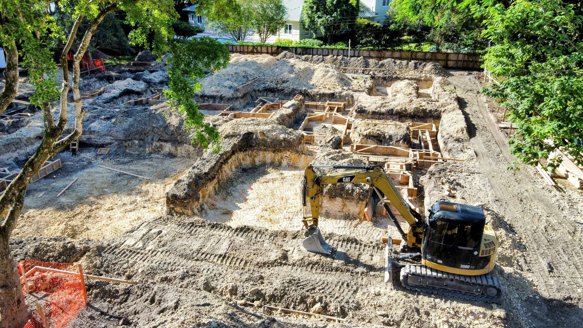 Excavator at a construction site, digging in a rectangular area surrounded by trees and debris.