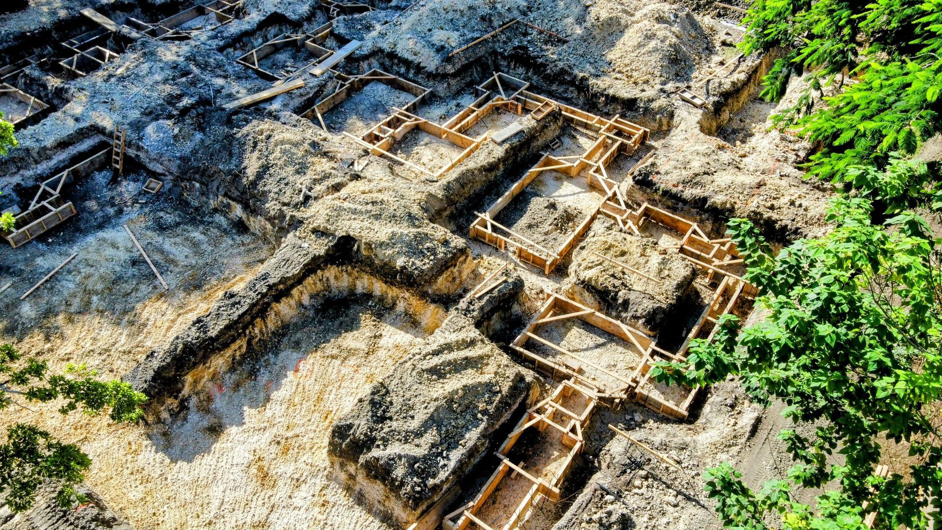 Aerial view of an archaeological dig site. Excavated building foundations with surrounding soil. Green foliage in the corner.