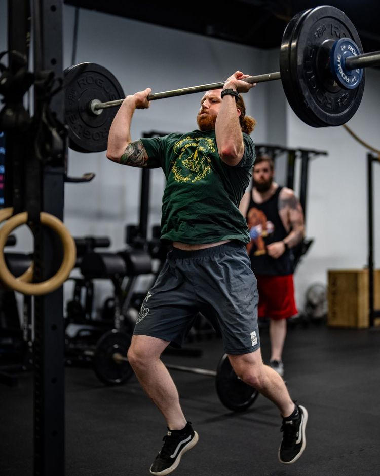 Athlete mid-air pressing a barbell overhead in a CrossFit gym, with another person in the background