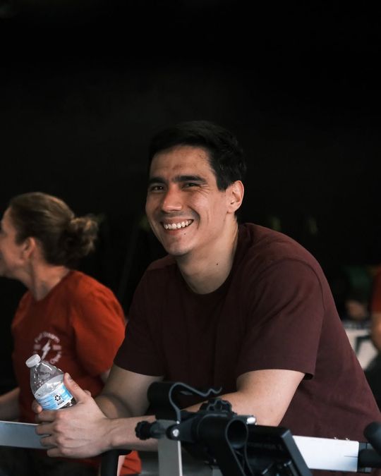 Smiling man in a maroon shirt holding a water bottle at a table, with a dark background.