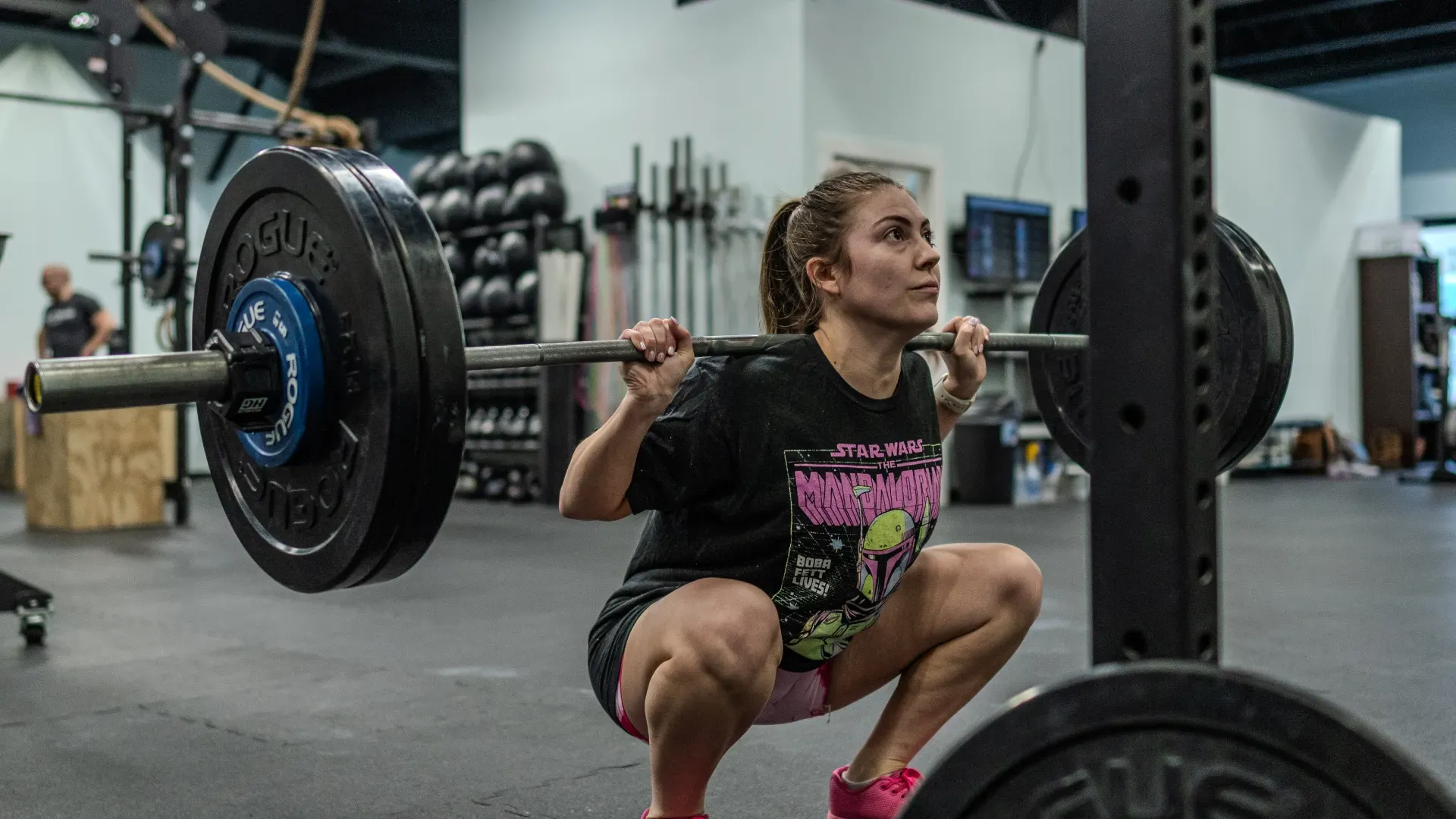 Woman performing a barbell squat in a gym, wearing a black shirt and pink shoes.