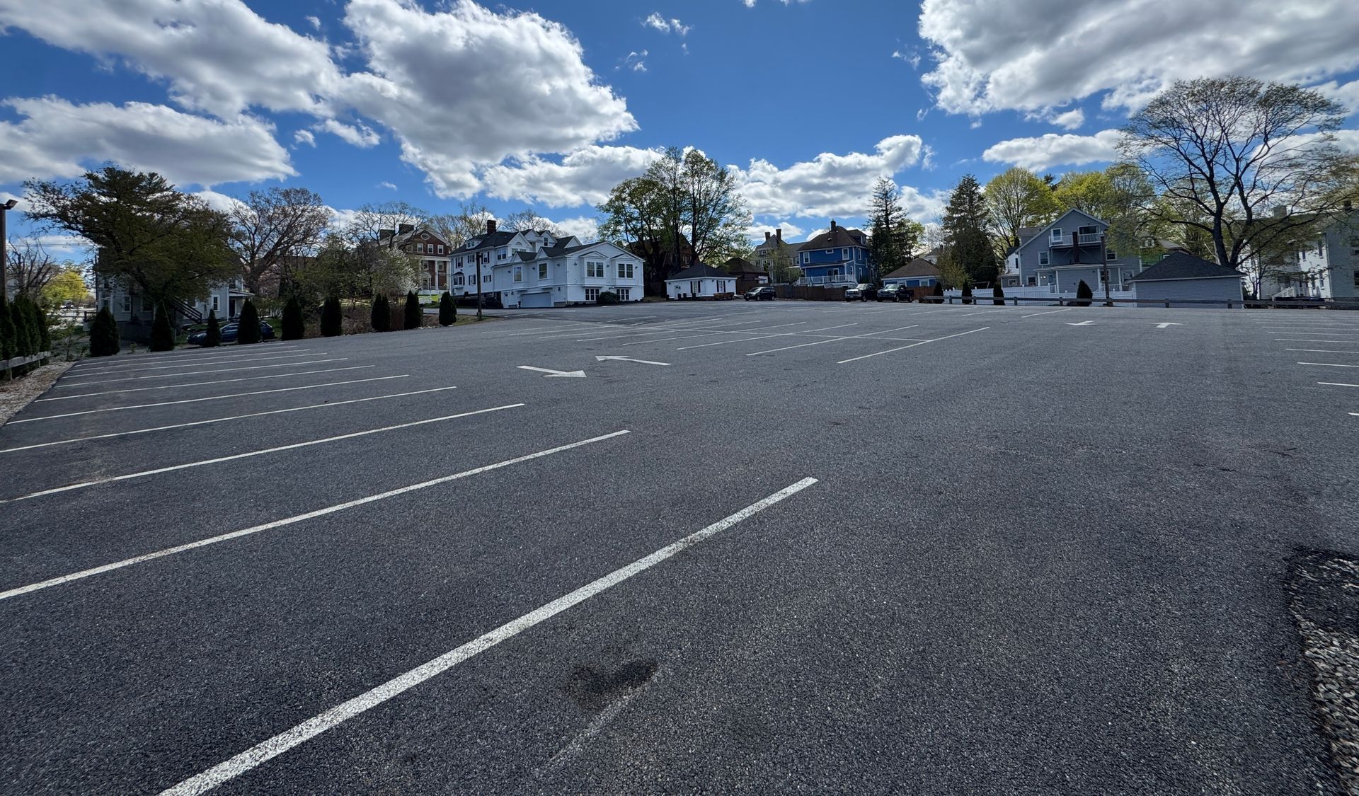 Empty parking lot in front of a white building with windows. Trees and sky visible.