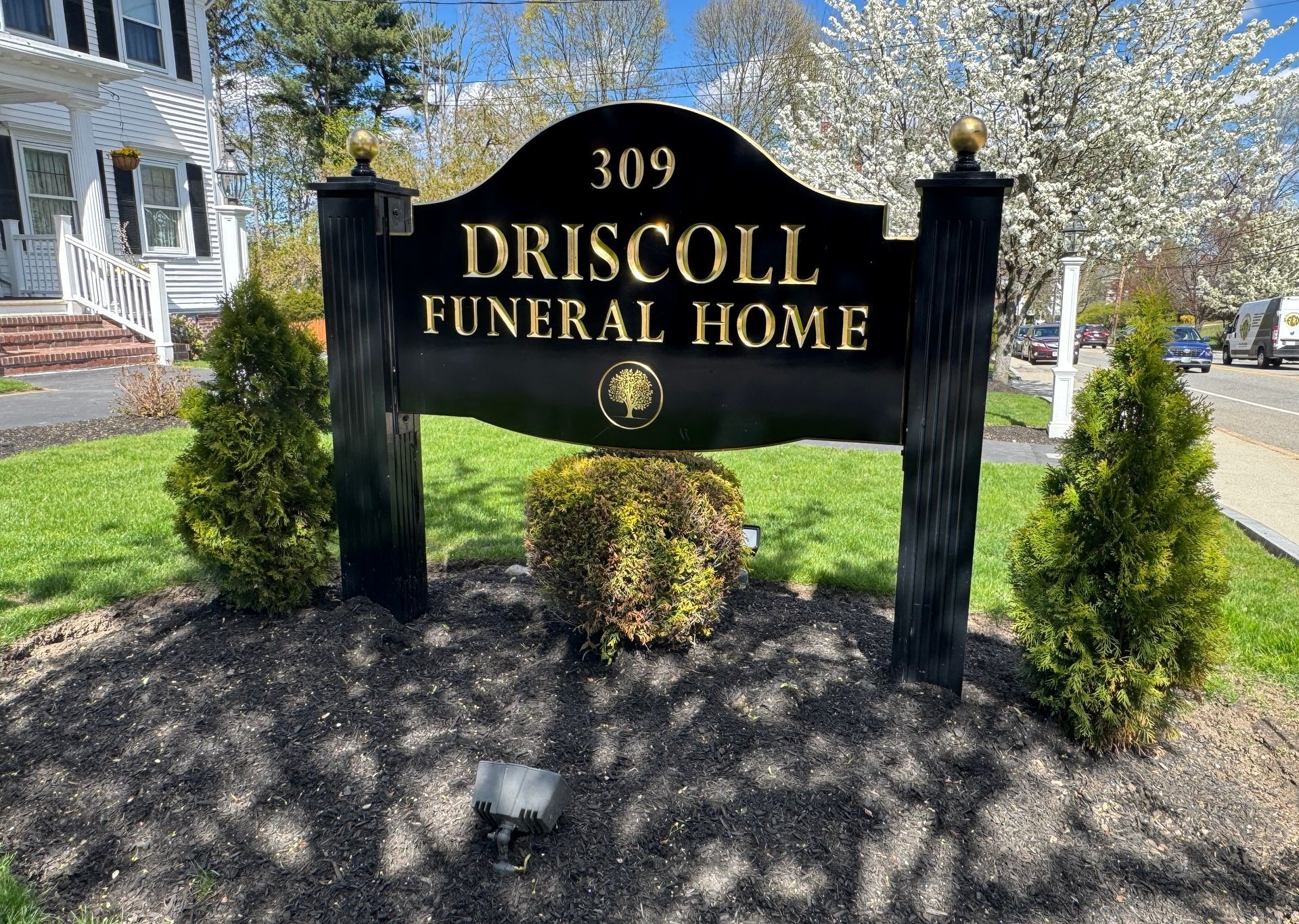 Sign for Driscoll Funeral Home, black with gold trim, in front of building with manicured landscaping.