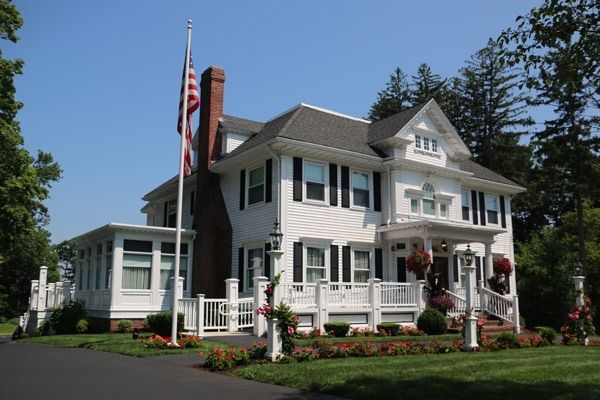 White two-story house with black shutters, a porch, and an American flag on a sunny day.