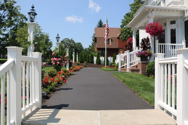 A black asphalt driveway leads to a brick building. White railing and flowers line the path under a blue sky.