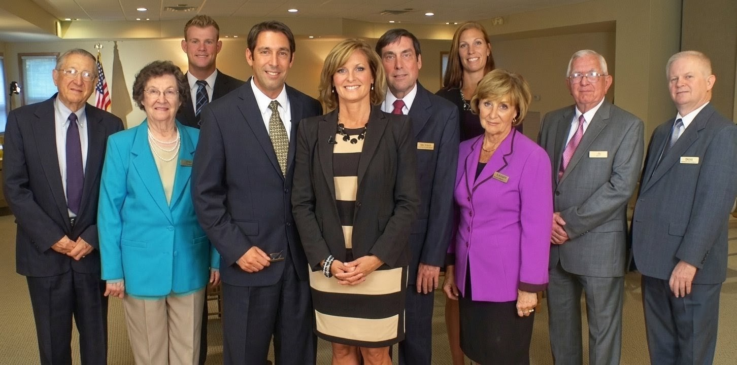 A group of nine people in professional business attire pose for a formal group portrait in a brightly lit indoor space.