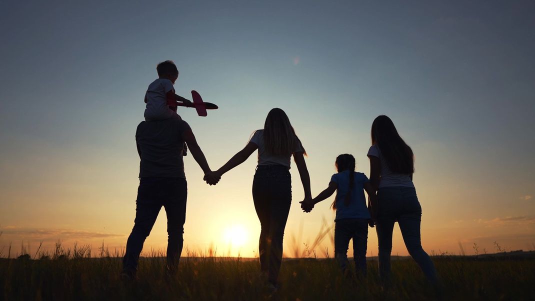 A family of five, including one child riding on shoulders, holding hands while walking through a field at sunset.
