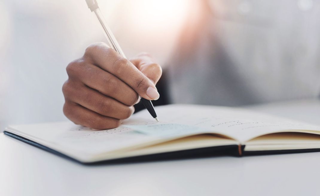 A hand holding a pen, writing in an open notebook on a white surface with soft lighting.