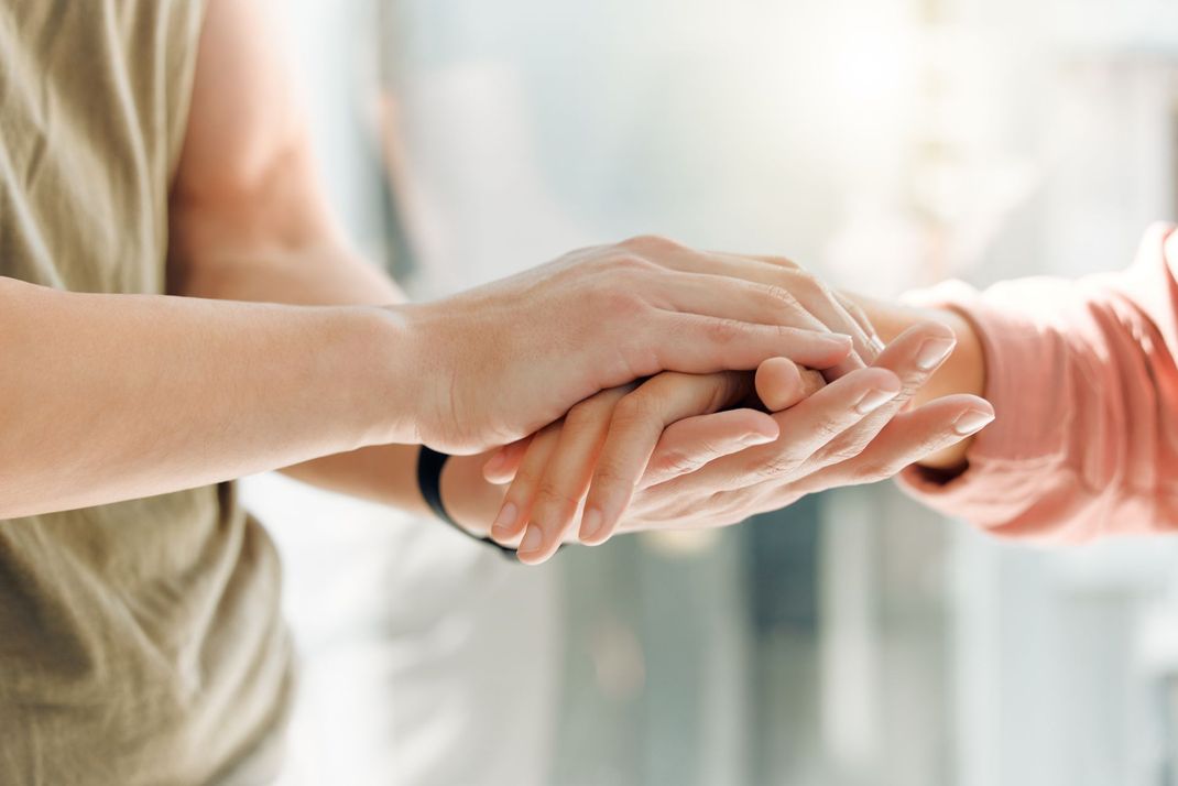 Two people hold hands in a gesture of support or comfort, with soft, bright natural lighting in the background.