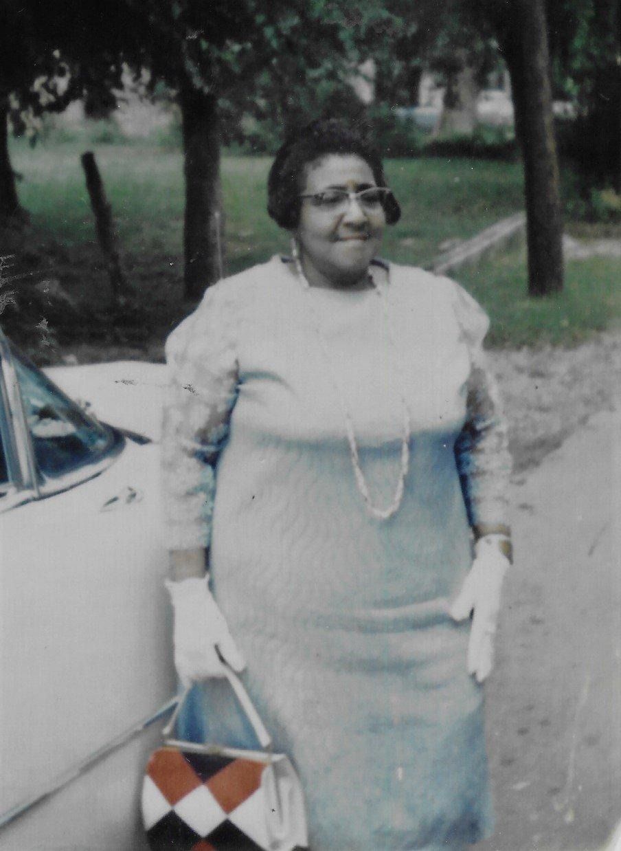 Woman in a light dress standing beside a car on a tree-lined path, holding a patterned handbag.